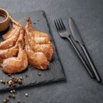 Delicacies. Grilled BBQ quail legs in marinade and spices on black slate with sauce
